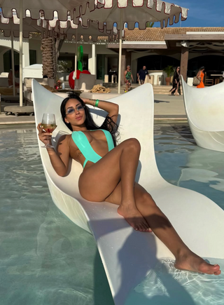 A woman reclining in a white curved pool float chair by a swimming pool, wearing a bright turquoise one-piece swimsuit and holding a glass of white wine.